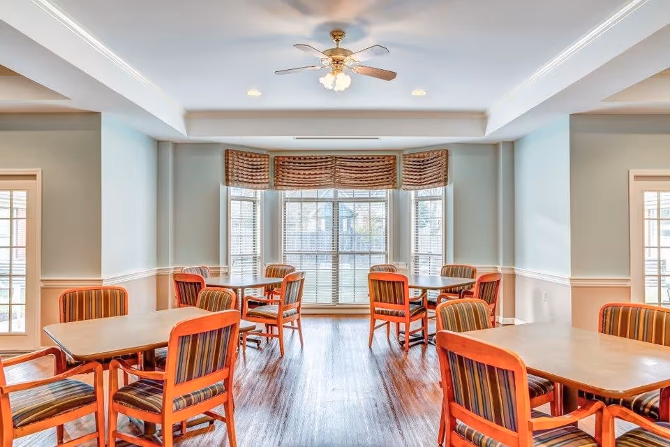 A bright dining room with several wooden tables and chairs featuring striped upholstery. Large windows with blinds and valances allow natural light to fill the room. The ceiling has recessed lighting and a ceiling fan with lights. The floor is wooden, and the walls are painted in light colors with white trim.