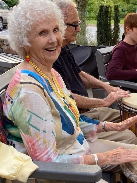 An elderly woman with curly white hair and colorful clothing is smiling while sitting outdoors with other people. She is wearing a yellow beaded necklace and appears to be participating in a group activity involving drums. Behind her, an elderly man and a younger person are also seated and engaged in the activity. The setting includes greenery and outdoor furniture.