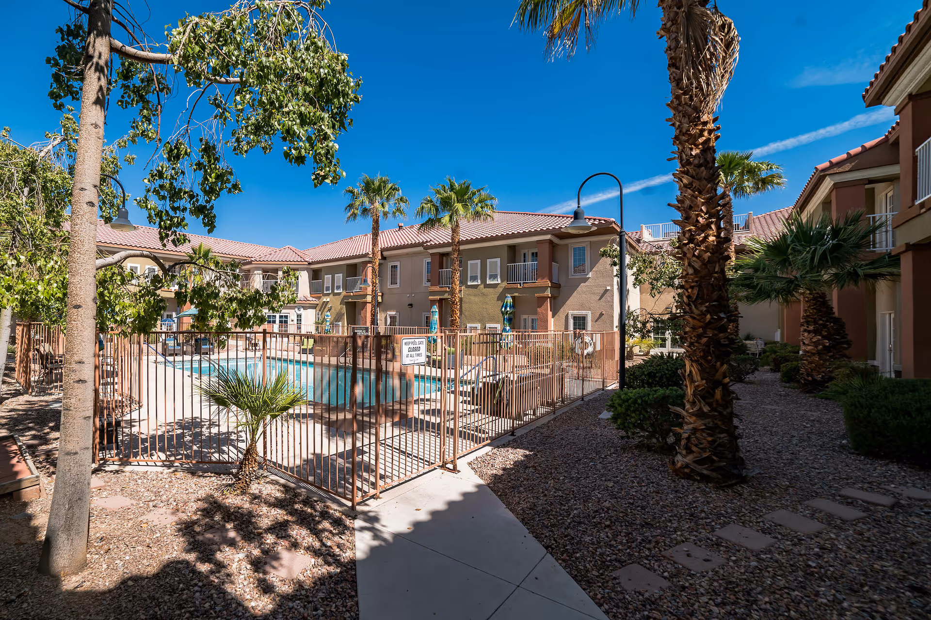 Outdoor view of a senior living facility courtyard at Acacia Springs featuring a fenced swimming pool surrounded by lounge chairs, palm trees, and a clear blue sky. The two-story building with balconies and tiled roofs encloses the courtyard.