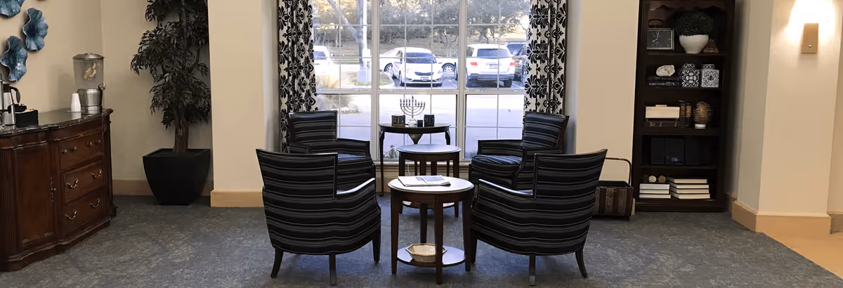 A seating area with four striped armchairs arranged around two small round wooden tables in front of a large window with patterned curtains. Outside the window, several parked cars are visible. To the left, there is a wooden cabinet with a water dispenser and cups, and a tall potted plant. To the right, there is a bookshelf with decorative items and books.