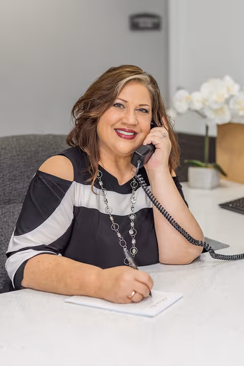 A smiling woman with shoulder-length brown hair is sitting at a white desk, talking on a corded phone while writing on a notepad. She is wearing a black and white top with cut-out shoulders and a silver necklace. Behind her, there is a blurred background with a white orchid plant and a computer keyboard.