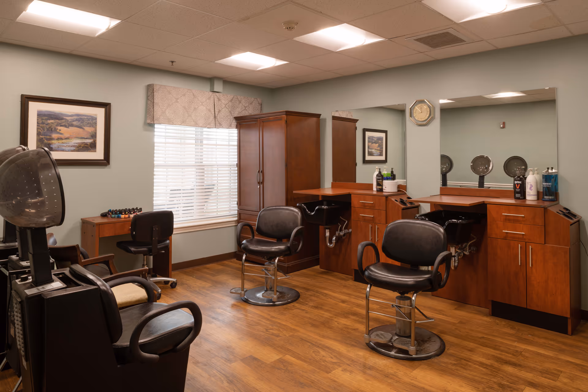 Interior view of a salon area in a senior living facility with two black salon chairs in front of wooden cabinets and mirrors. There is a hair dryer chair on the left side, a window with blinds and a valance, a framed landscape picture on the wall, and a small desk with a chair and nail polish bottles.