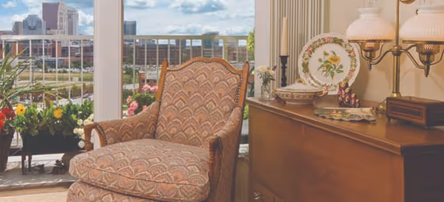 Upholstered armchair next to a wooden sideboard with decorative plates and a lamp, facing sliding glass doors opening to a balcony with flowers and a city skyline view.