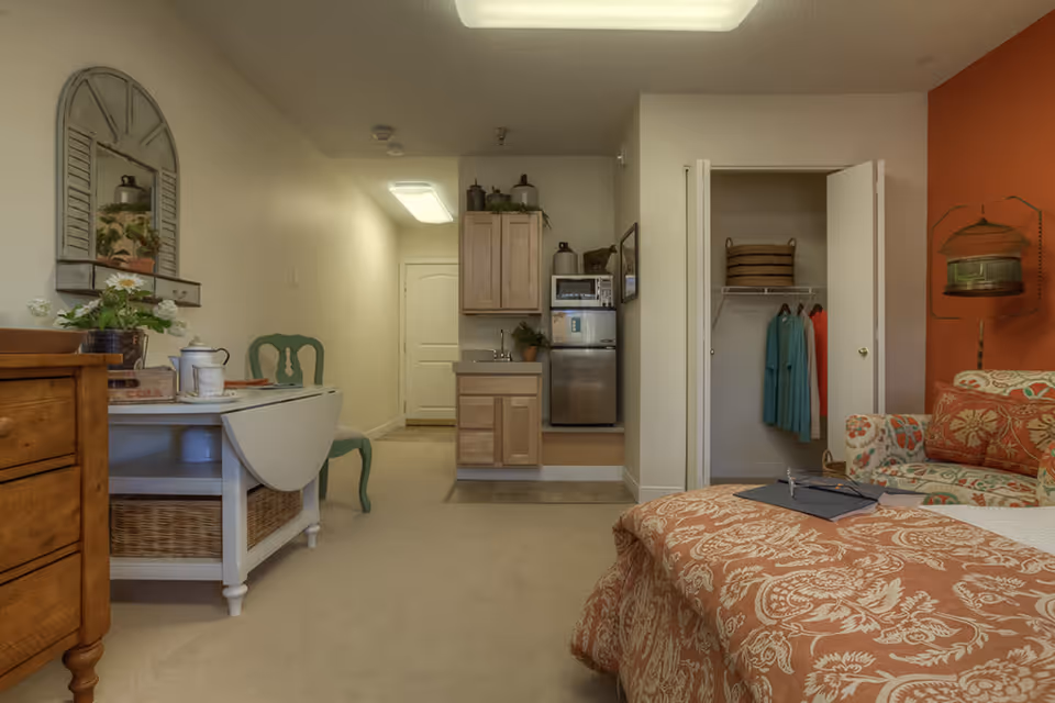 A cozy studio apartment interior with a bed covered in an orange patterned bedspread, a floral armchair, an open closet with clothes hanging, a small kitchenette with a mini fridge and microwave, a green chair, and a white table with a basket underneath. The walls are painted beige with one accent wall in orange.