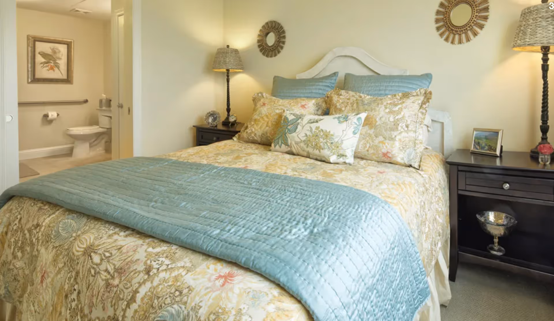 A cozy bedroom with a made bed dressed in floral bedding and a teal quilt, nightstands with lamps, and an open doorway revealing an adjacent bathroom.