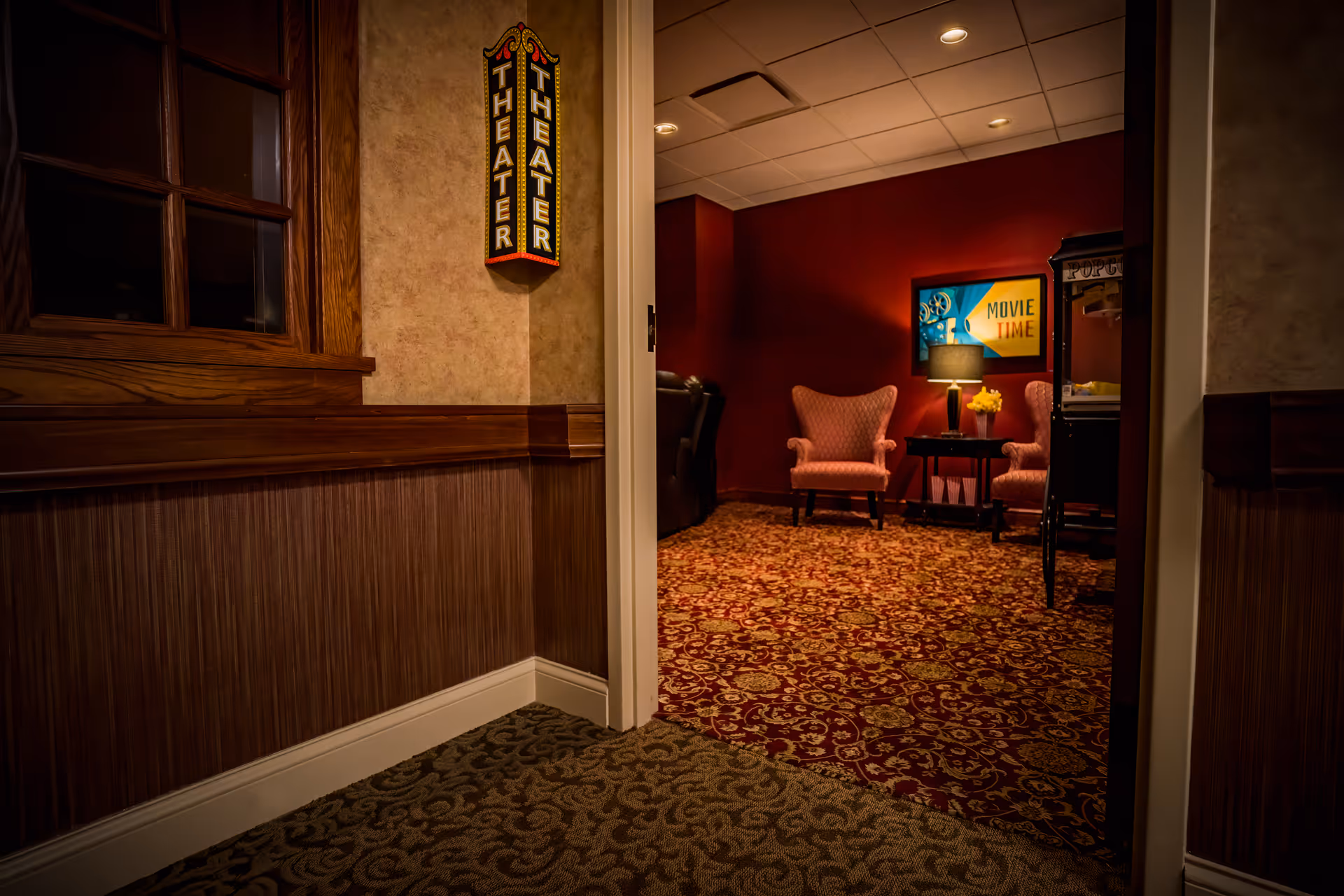 View through an open doorway into a cozy theater room with patterned carpet, two upholstered armchairs, a side table with a lamp and flowers, a popcorn machine, and a colorful 'Movie Time' sign on the wall. Outside the doorway, there is a hallway with wood paneling and a 'THEATER' sign on the wall.