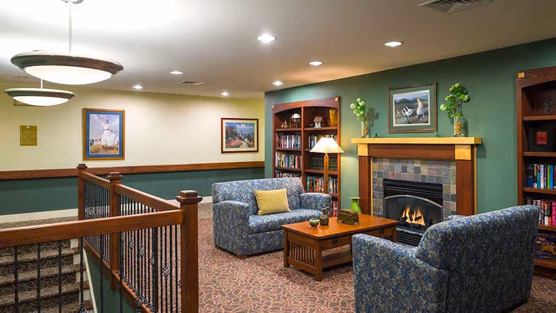 A cozy senior living common area with two blue patterned armchairs and a matching loveseat arranged around a wooden coffee table in front of a lit fireplace with a tiled surround. The walls are painted green with wooden trim, and there are bookshelves filled with books on either side of the fireplace. Framed artwork and decorative plants are displayed above the fireplace and on the walls. A staircase with wooden railing and iron balusters is visible on the left side.