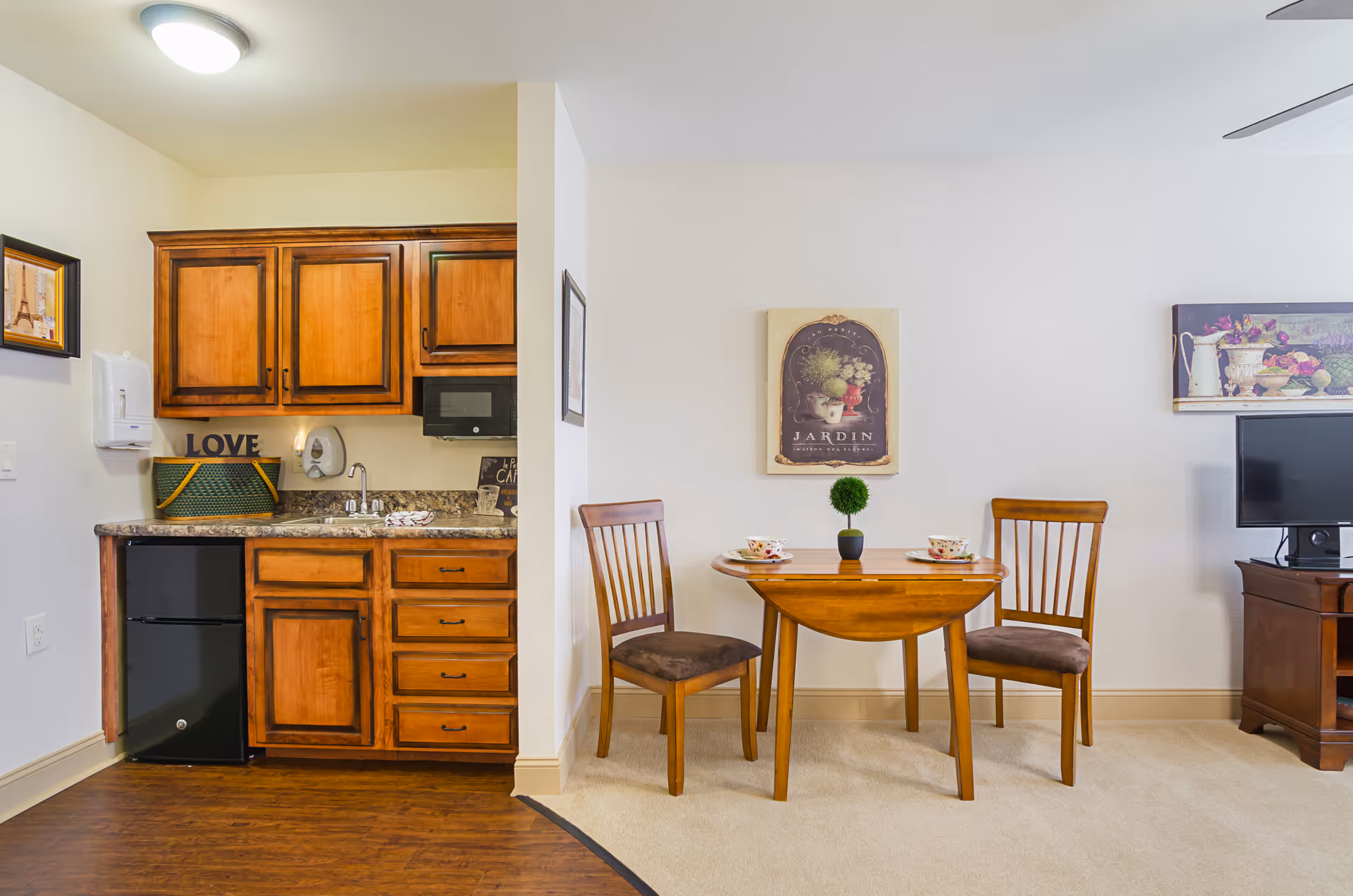 A cozy assisted living apartment interior featuring a small kitchenette with wooden cabinets, a mini refrigerator, a sink, and a microwave. Adjacent to the kitchenette is a wooden dining table set with two chairs, a small potted plant, and two teacups. The room has light-colored walls adorned with framed artwork and a TV on a wooden stand.