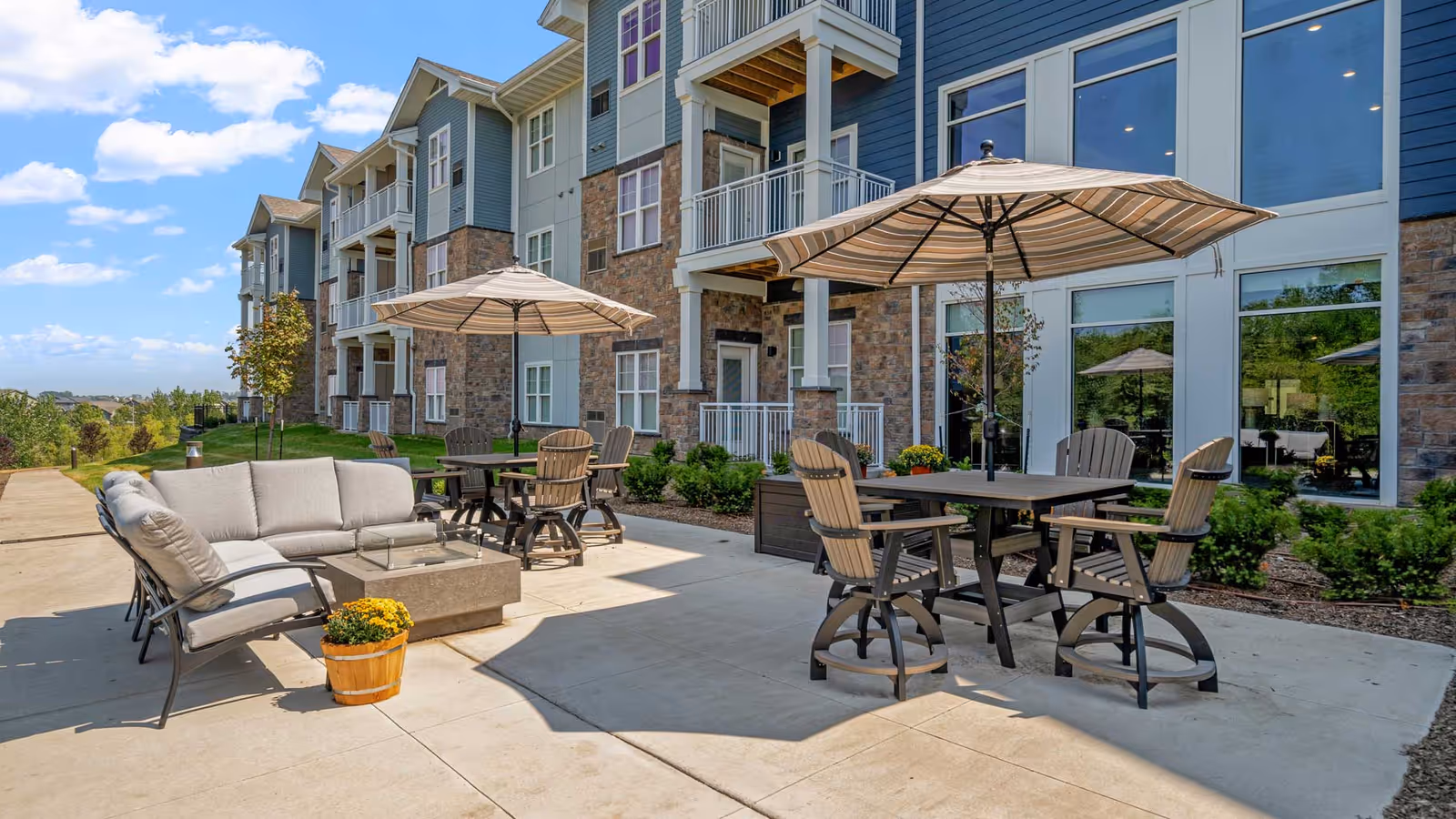 Outdoor patio with tables, umbrellas, lounge chairs and potted flowers outside a multi-story senior living building.