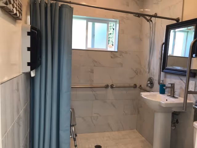 A bathroom with a walk-in shower featuring a blue shower curtain, grab bars on the tiled walls, a small window, a pedestal sink with a soap dispenser, a mirror above the sink, and a paper towel dispenser on the wall.