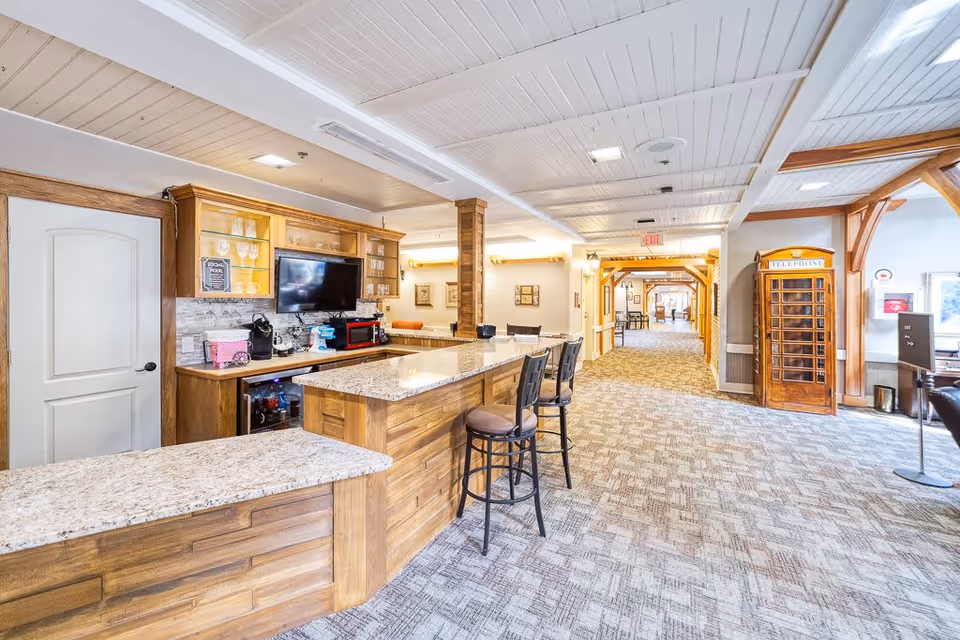Interior view of a senior living facility common area with a wooden counter and bar stools, a mounted TV, glass cabinets, and a vintage-style wooden telephone booth. The space has carpeted floors, white paneled ceilings, and wooden beams, with a hallway leading to other rooms.