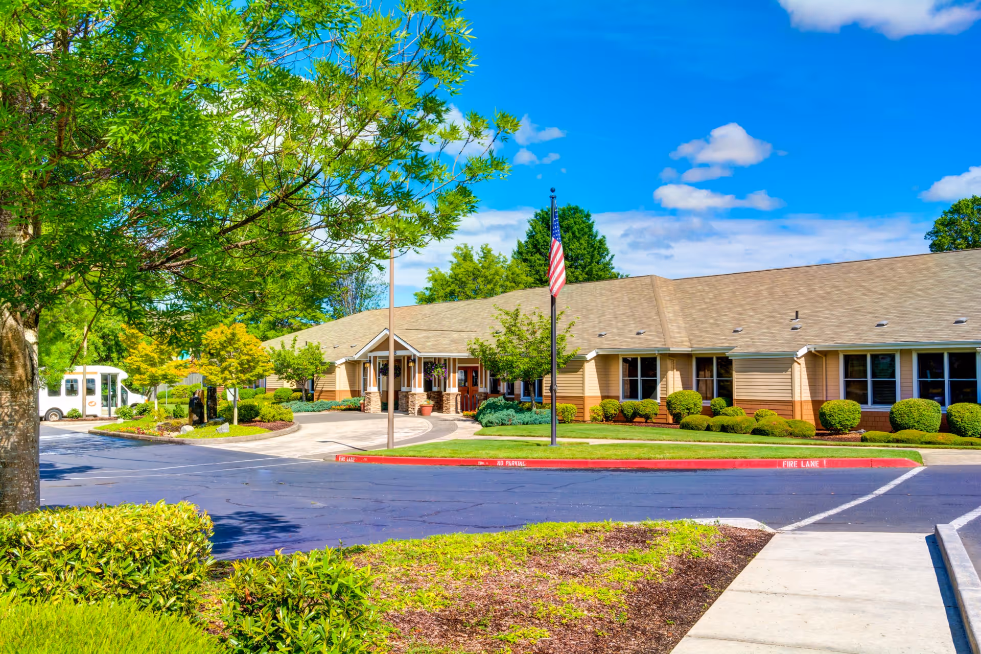 Exterior view of Monterey Court Memory Care facility with a single-story building, neatly trimmed bushes, trees, a flagpole with an American flag, and a clear blue sky with some clouds.