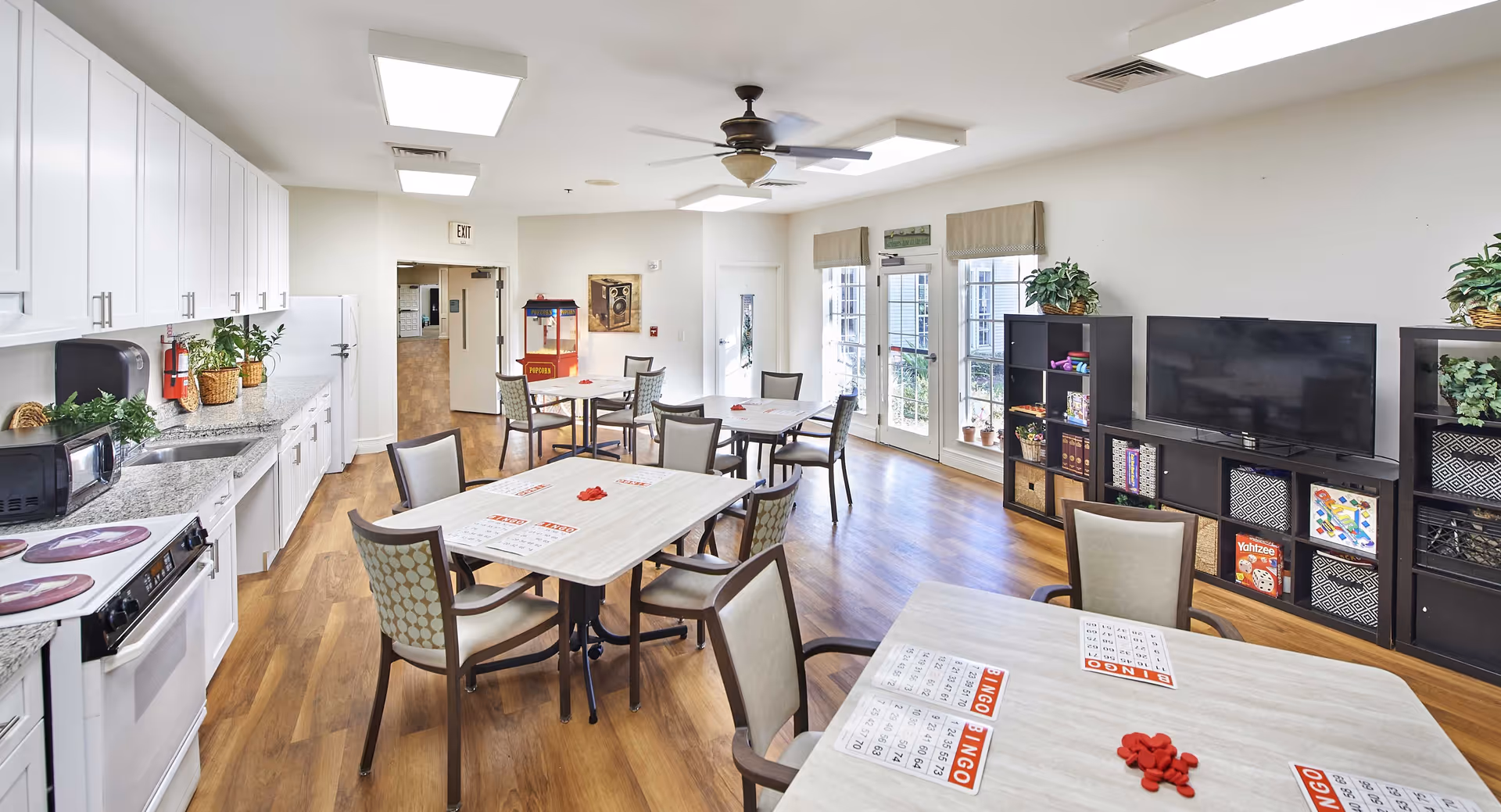 A bright and spacious common room with a kitchenette on the left side featuring white cabinets, a microwave, stove, and sink. Several tables and chairs are arranged in the center of the room, each table set up with bingo cards and red bingo chips. On the right side, there is a TV on a black shelving unit filled with board games and plants. Large windows and a glass door let in natural light, and a ceiling fan is mounted above.