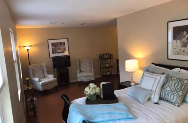 A cozy bedroom with a neatly made bed featuring multiple pillows and a light blue throw blanket. There is a bedside table with a lamp and a framed picture on the wall above the bed. In the background, two upholstered armchairs are positioned near a small TV on a stand, with a floor lamp providing additional lighting. The room has warm beige walls and wooden flooring.