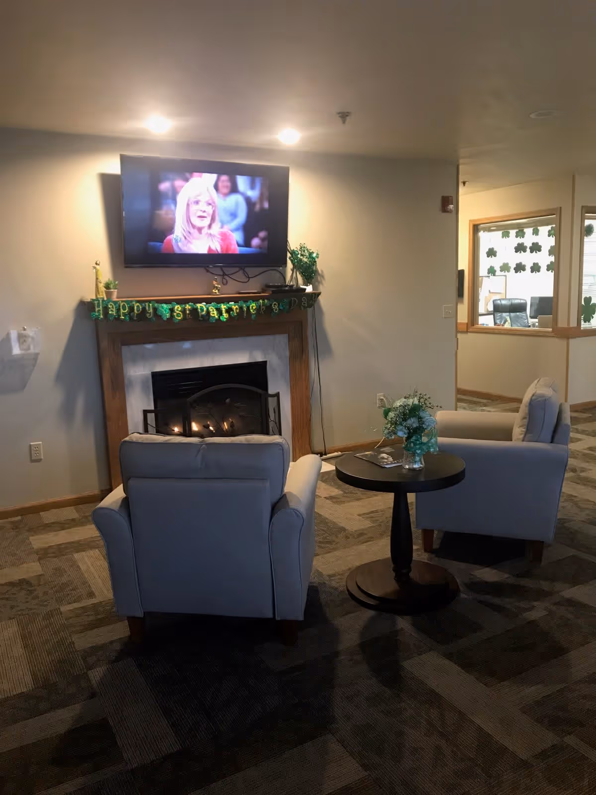 A cozy living room area with two beige armchairs facing a fireplace with a wooden mantel. Above the mantel is a flat-screen TV mounted on the wall showing a woman with blonde hair. The mantel is decorated with a green garland that reads 'Happy St. Patrick's Day' and some small plants. A round wooden side table with a vase of flowers and a magazine is placed between the chairs. The room has patterned carpet flooring and light-colored walls. In the background, there is a window decorated with shamrock stickers, revealing an office space.