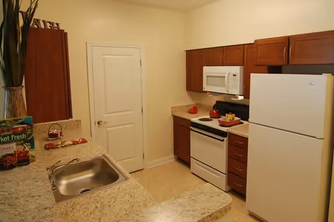 A compact kitchen with beige walls and granite countertops. The kitchen features a white refrigerator, white microwave, and white stove with a red kettle on it. There are wooden cabinets above and below the countertops. A stainless steel sink is visible on the left side, along with some kitchen items including a box of frozen food and a small basket. A closed white door is in the background.