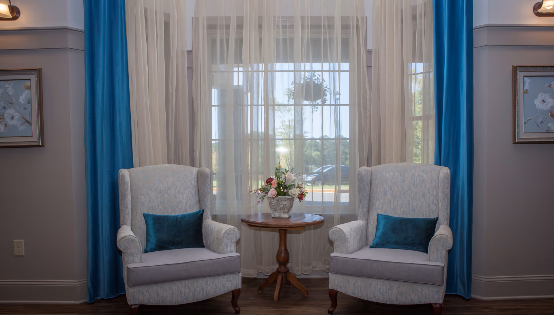 Two upholstered armchairs with teal accent pillows are positioned on either side of a small round wooden table holding a floral arrangement. Behind them is a large window with sheer white curtains and teal drapes, allowing natural light to fill the room. The walls are decorated with framed floral artwork.