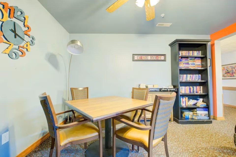 A small game room with a square wooden table surrounded by four chairs with yellow cushions. A tall floor lamp stands in the corner, and a colorful clock is mounted on the wall. A black bookshelf filled with books is against the wall, next to a small cabinet labeled for donated books and book returns. The room has light-colored walls and carpeted flooring.