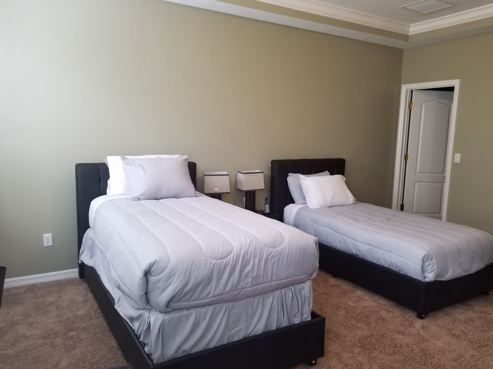 A bedroom with two single beds, each with white bedding and pillows. Between the beds is a small nightstand with two lamps and a telephone. The walls are painted light green and there is a white door on the right side of the room.