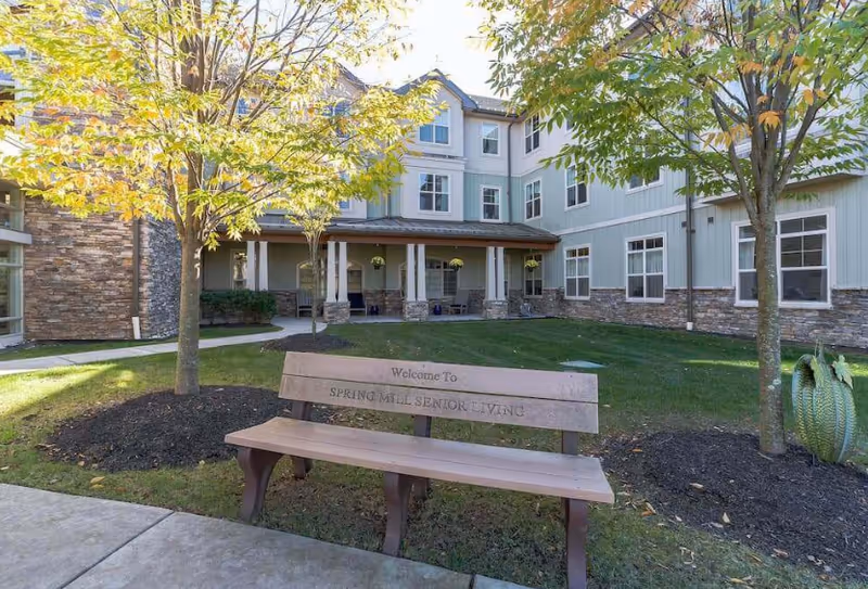 Outdoor courtyard area of a senior living facility with a wooden bench that reads 'Welcome To Spring Mill Senior Living', surrounded by green grass, trees with autumn leaves, and a multi-story building with stone and light-colored siding in the background.