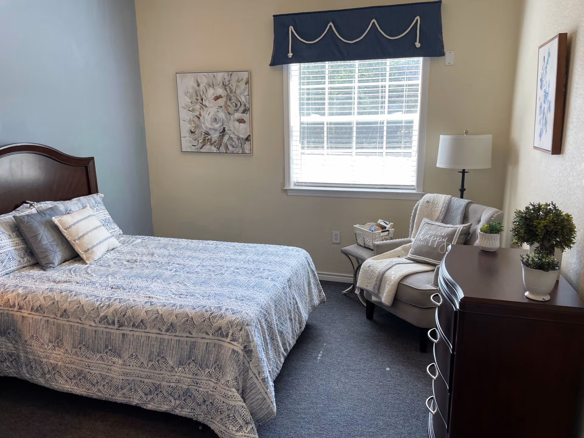 A bedroom with a neatly made bed featuring patterned bedding and several pillows. There is a wooden headboard, a window with blinds and a blue valance, a cushioned armchair with a throw blanket and a decorative pillow, a floor lamp, a wooden dresser with two small potted plants, and framed floral artwork on the walls.