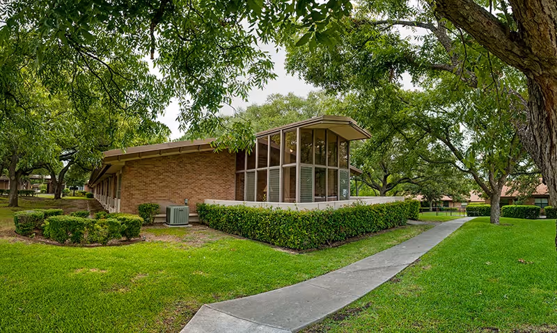 Exterior view of a single-story brick building with large windows and a slanted roof, surrounded by green grass, trimmed bushes, and mature trees. A concrete pathway curves alongside the building through the landscaped grounds.
