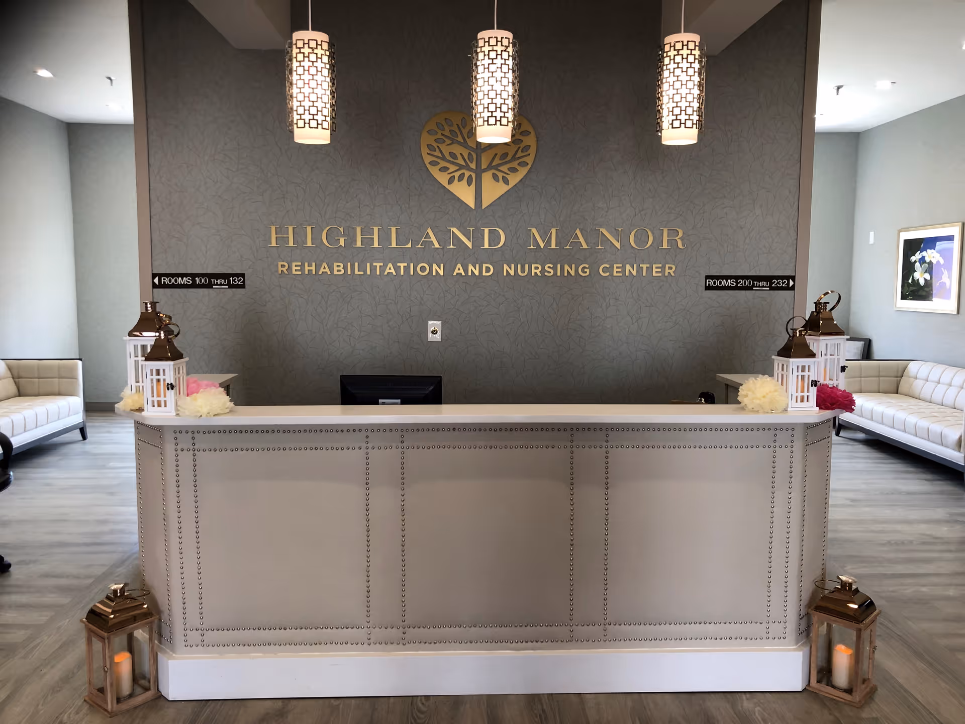 Reception desk area of Highland Manor Rehabilitation and Nursing Center with a decorative wall sign, three hanging pendant lights, two white sofas on either side, and lanterns with candles placed on the floor and desk.