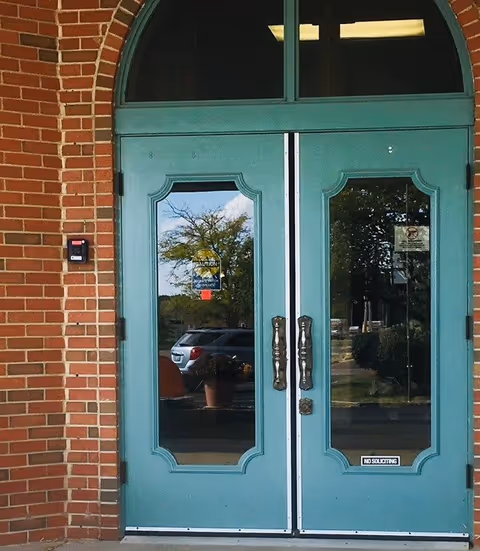 Double teal-colored entrance doors with glass panels set in a brick wall. The glass panels reflect a parking lot with cars and trees. There are signs on the doors including a 'No Soliciting' sign and a notice about a security system.