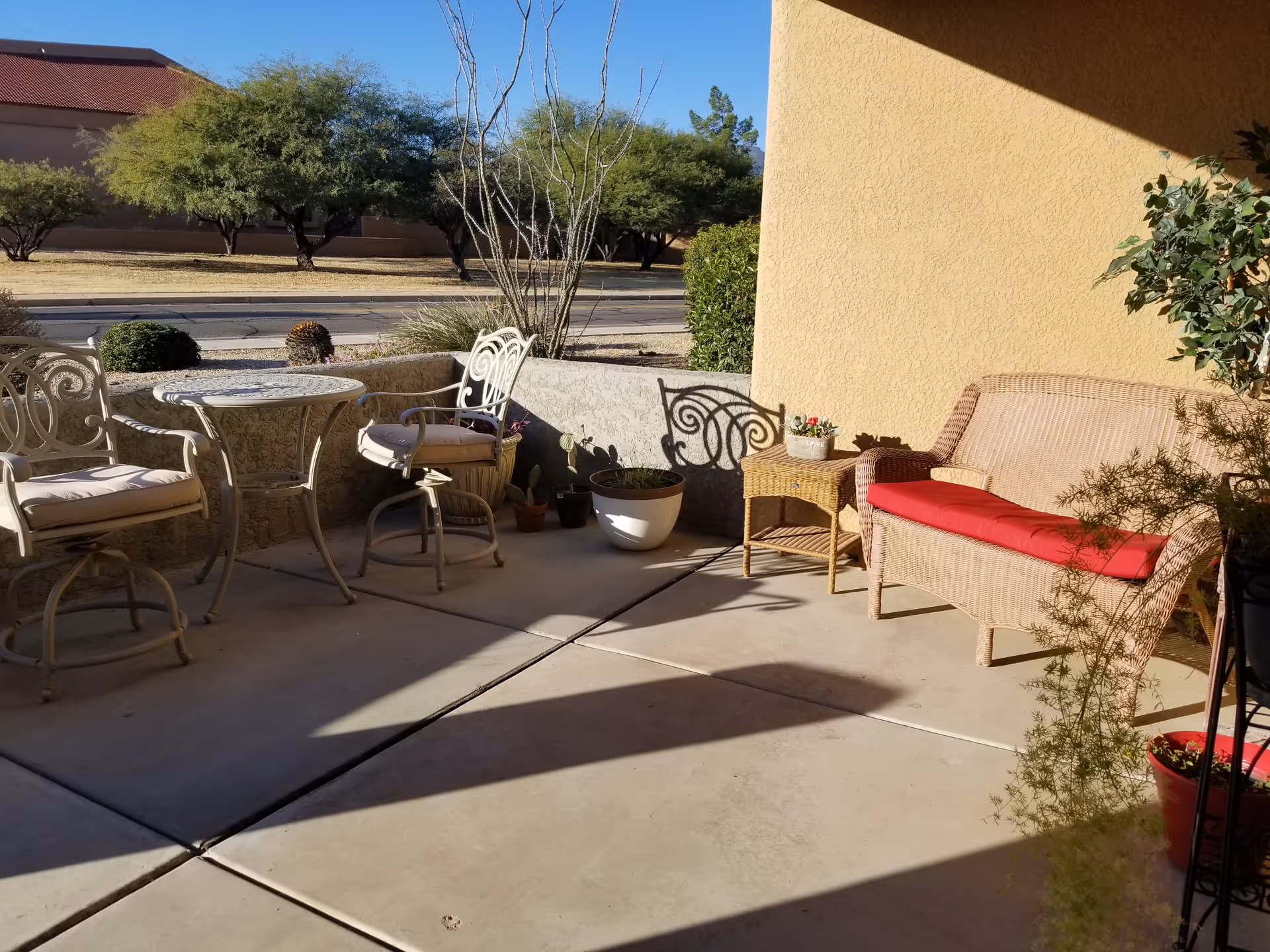 Outdoor patio area with a small round table and two cushioned chairs on the left, a wicker loveseat with a red cushion on the right, a small wicker side table with a potted plant, and several other potted plants around. Trees and a road are visible in the background under a clear blue sky.
