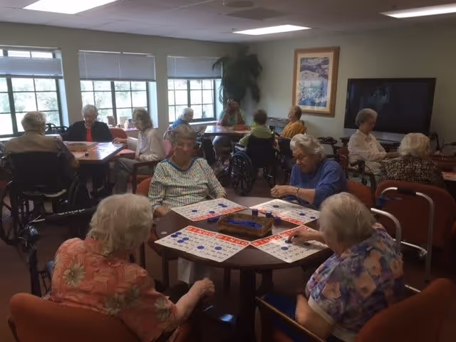 A group of elderly women sitting around tables in a well-lit room playing bingo. The room has large windows with blinds, a large TV on the wall, and framed artwork. Some women are in wheelchairs, and others are seated in chairs, all engaged in the game.