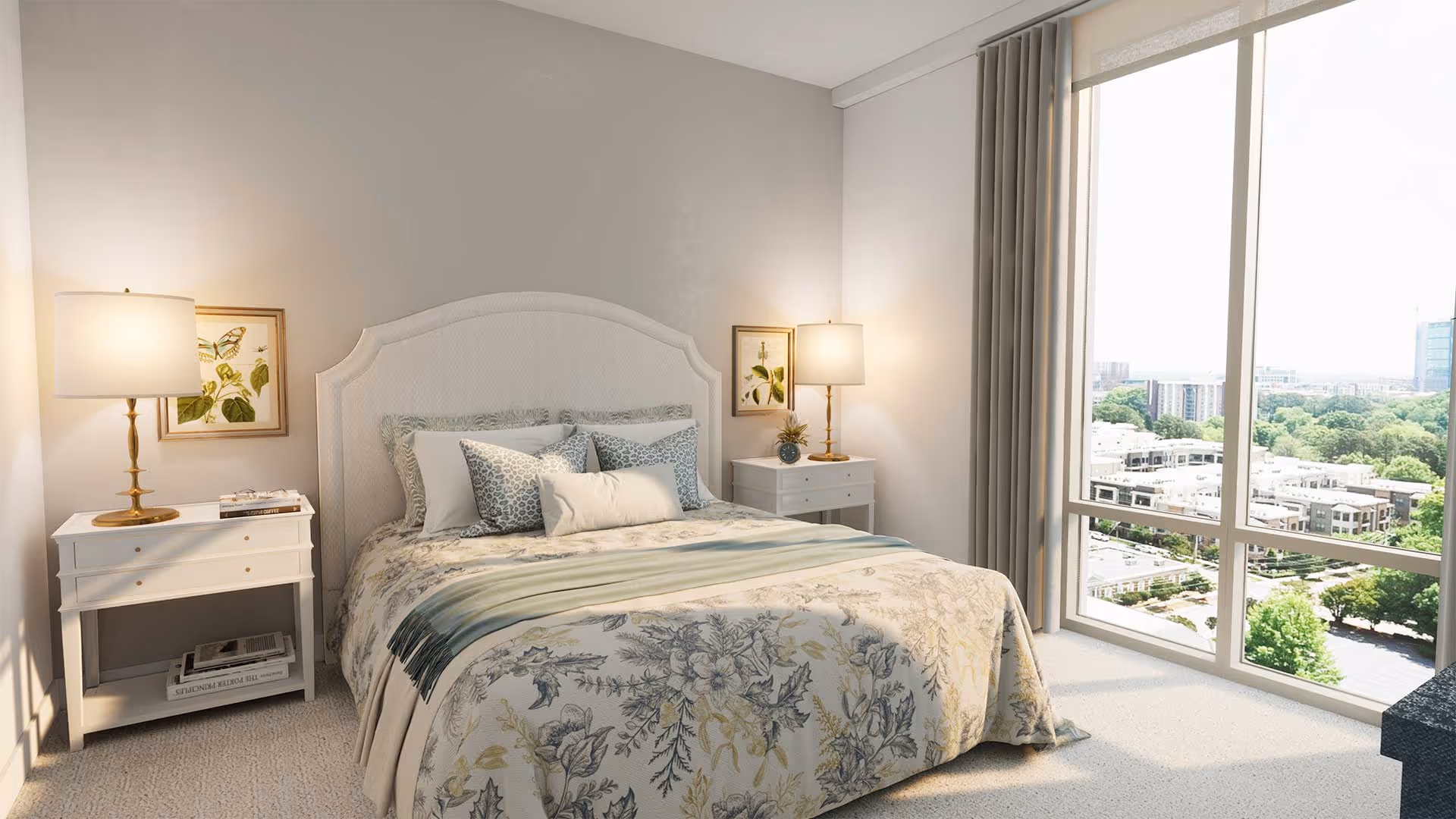 A bright bedroom with a large window offering a city view. The room features a bed with floral bedding and multiple pillows, two white nightstands each with a lamp and framed botanical artwork above them. Light-colored curtains frame the window.