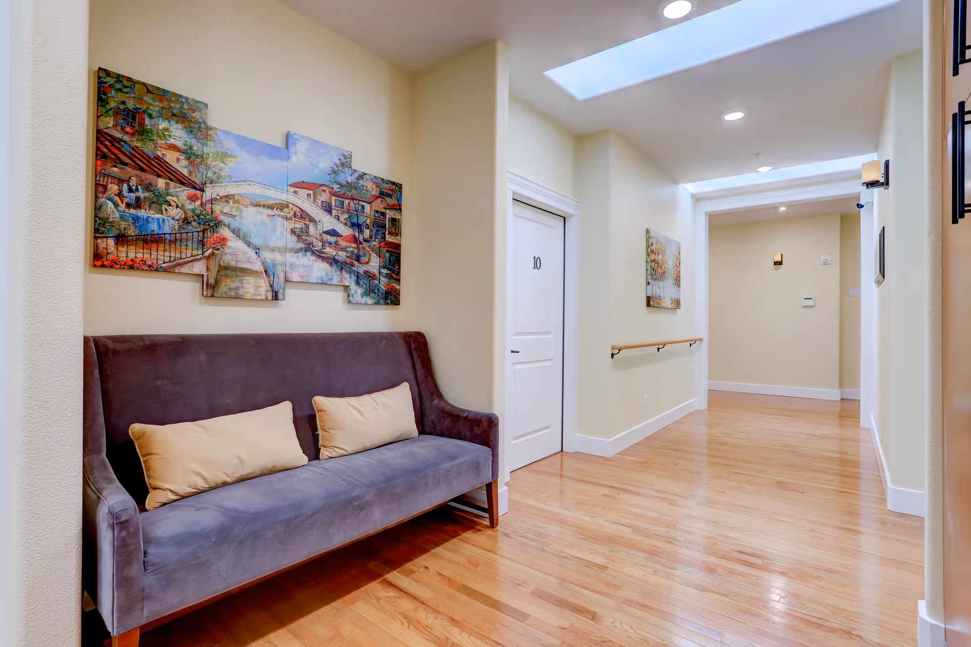 A well-lit hallway with light yellow walls and wooden flooring. On the left side, there is a dark gray upholstered bench with two beige cushions beneath a colorful multi-panel painting of a canal scene. The hallway features white doors, one marked with the number 10, and handrails along the walls. Skylights and recessed lighting illuminate the space.
