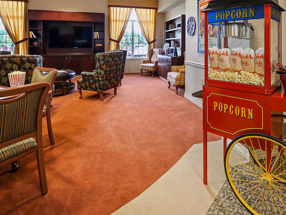 A cozy living room area with patterned armchairs, a large flat-screen TV mounted on a wooden entertainment center, and a vintage-style red popcorn machine filled with popcorn bags. The room has large windows with yellow curtains, a carpeted floor, and additional seating including striped chairs and a small desk with a chair in the background.