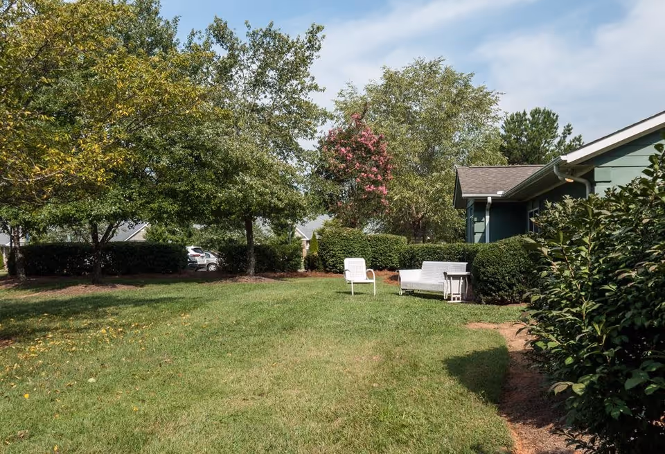 Outdoor grassy area with trees and bushes surrounding it. There are two white chairs and a white bench with a small table next to it on the grass. A green building is partially visible on the right side under a partly cloudy sky.