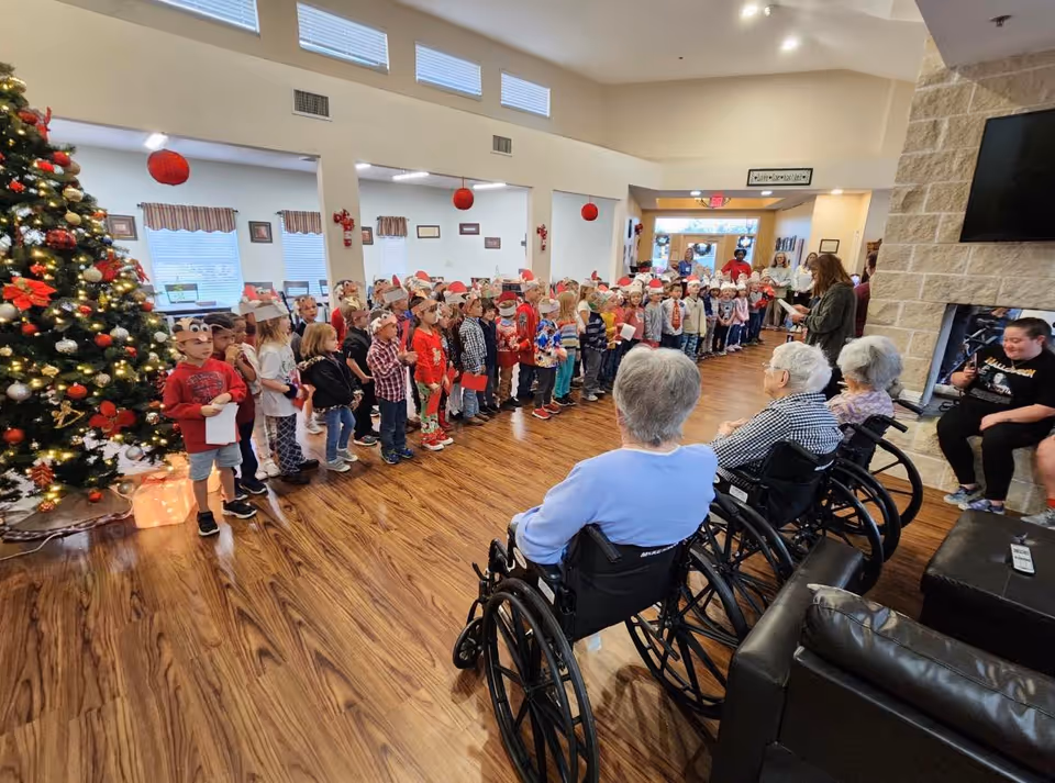 A group of children wearing festive holiday hats and holding papers stand in a line inside a senior living facility decorated for Christmas, with a large decorated Christmas tree on the left. Several elderly residents in wheelchairs and a few other adults are seated or standing nearby, watching the children. The room has wooden floors, a stone fireplace, and a TV mounted on the wall.