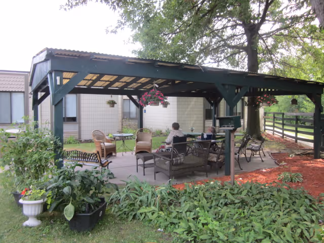 Outdoor covered patio area with seating including chairs and benches, surrounded by plants and trees. Two people are sitting and conversing under the pergola structure. The background shows a building and a fenced yard.