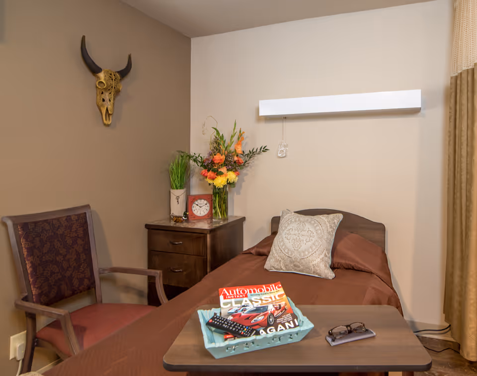 A cozy bedroom with a single bed covered in a brown blanket and a decorative pillow. Next to the bed is a wooden nightstand with a vase of colorful flowers, a small plant, and a red clock. A wooden chair with a patterned cushion is placed against the wall. Above the bed is a white wall-mounted light fixture, and a decorative bull skull hangs on the adjacent wall. A small table in front of the bed holds a magazine, a TV remote, and a pair of glasses.