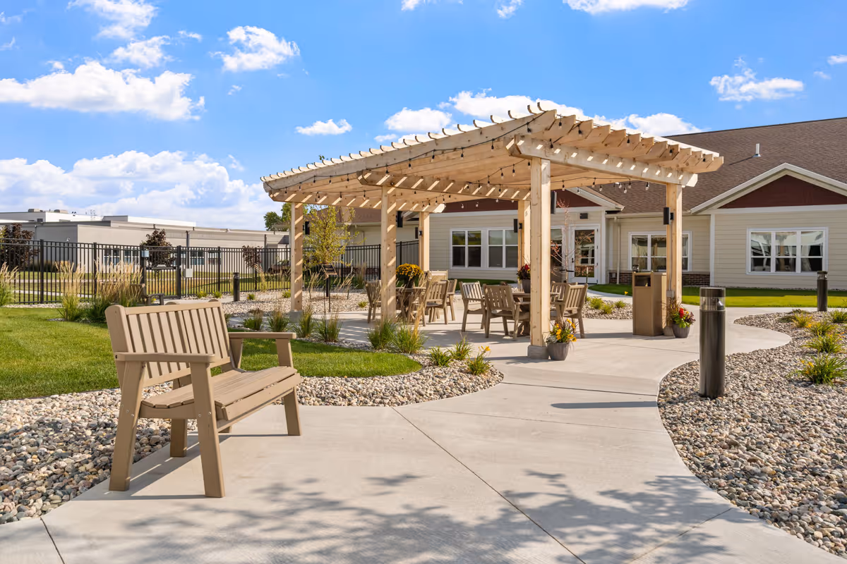 Outdoor seating area at Fargo Elim Health Care Center featuring a wooden pergola with string lights, several chairs and tables underneath, a bench along a curved concrete pathway, surrounded by landscaped grass and rocks, with the facility building in the background under a partly cloudy blue sky.