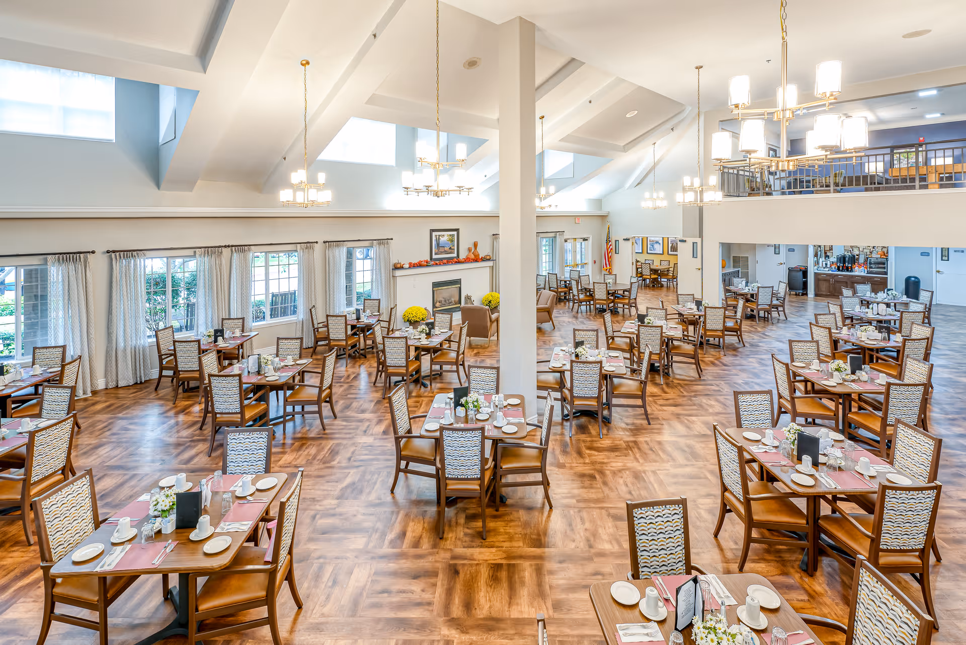 A spacious, well-lit dining room in a senior living facility with multiple wooden tables and chairs arranged neatly. Each table is set with white plates, cups, and silverware, along with small flower arrangements. Large windows with curtains allow natural light to fill the room, and there are chandeliers hanging from the ceiling. The room features a fireplace with decorative items and a second-floor balcony overlooking the dining area.