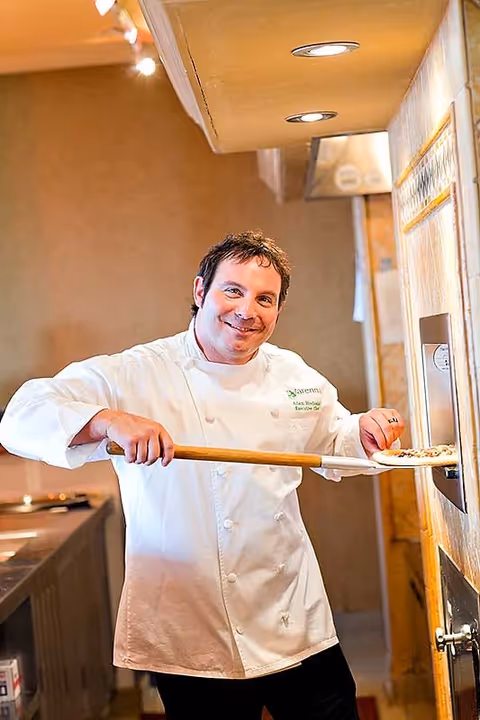 A chef wearing a white coat with the Varenna logo is smiling while using a wooden pizza peel to place or remove a pizza from a stone oven in a kitchen setting.
