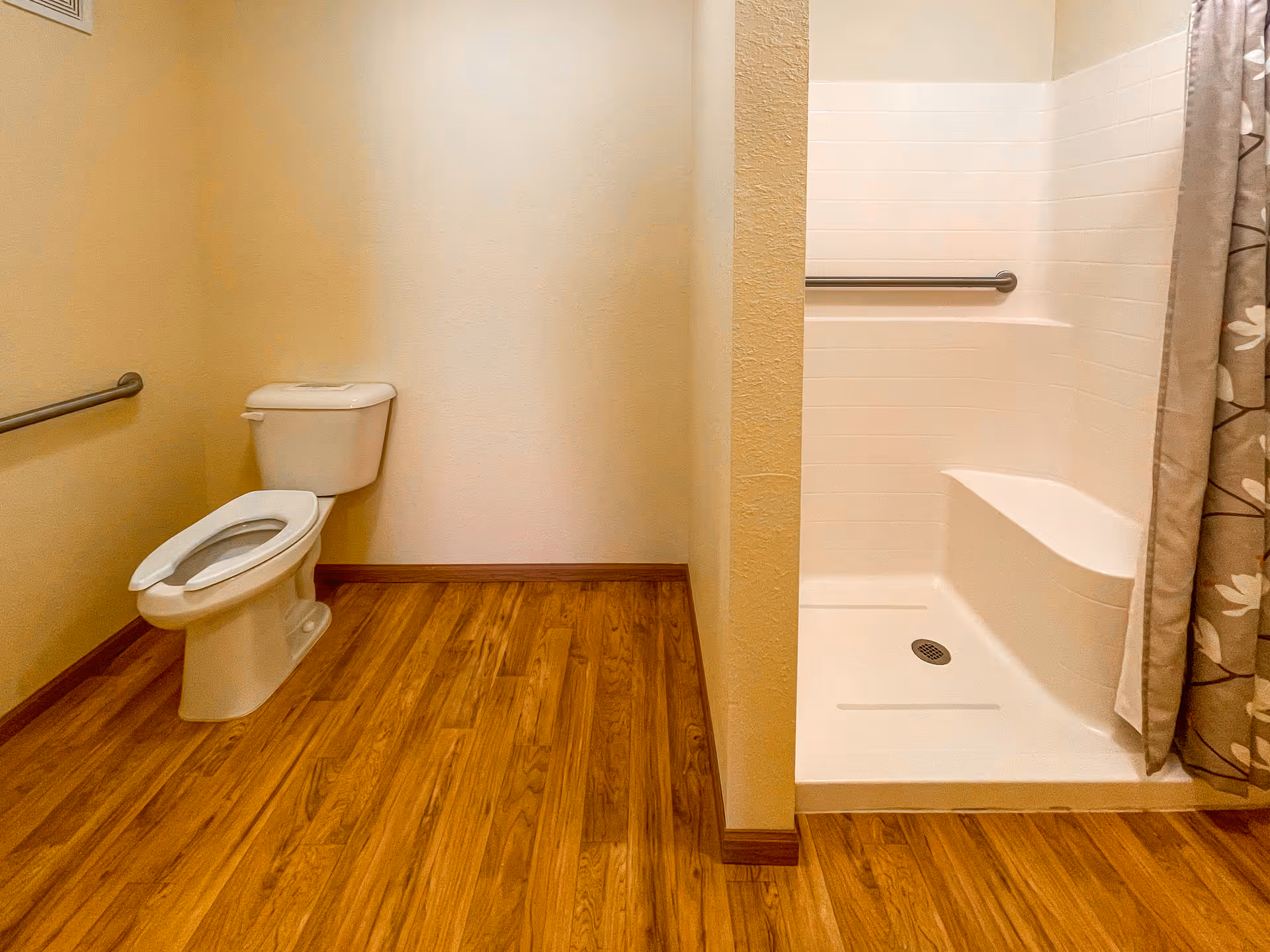 A bathroom with a white toilet on the left side and a walk-in shower with a built-in seat on the right side. The floor is wood-patterned, and there are grab bars installed near the toilet and inside the shower. A floral-patterned shower curtain is partially visible.