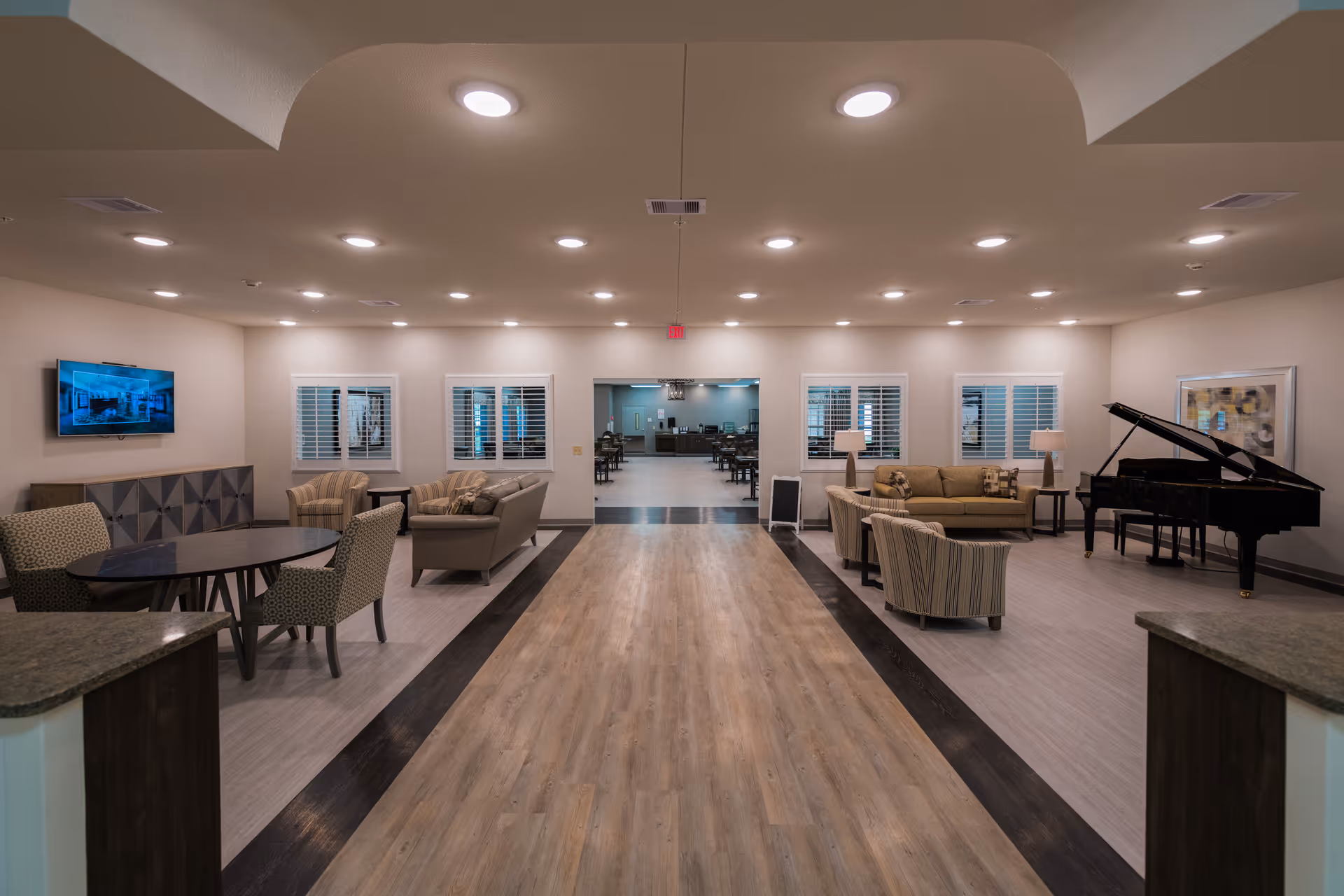 A spacious and well-lit common area in a senior living facility featuring multiple seating arrangements with armchairs and sofas. There is a round table with chairs on the left side, a flat-screen TV mounted on the wall, and a grand piano on the right side. The room has wood and tile flooring, recessed ceiling lights, and windows with white shutters. In the background, there is an open doorway leading to a dining area with tables and chairs.