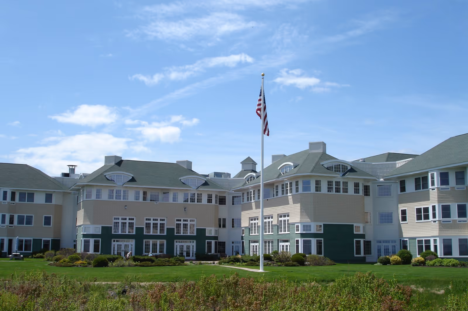 Exterior view of a large multi-story senior living facility building with green and beige siding, numerous windows, and a green roof under a blue sky with scattered clouds. A tall flagpole with an American flag stands in front of the building, surrounded by well-maintained green lawns and shrubs.