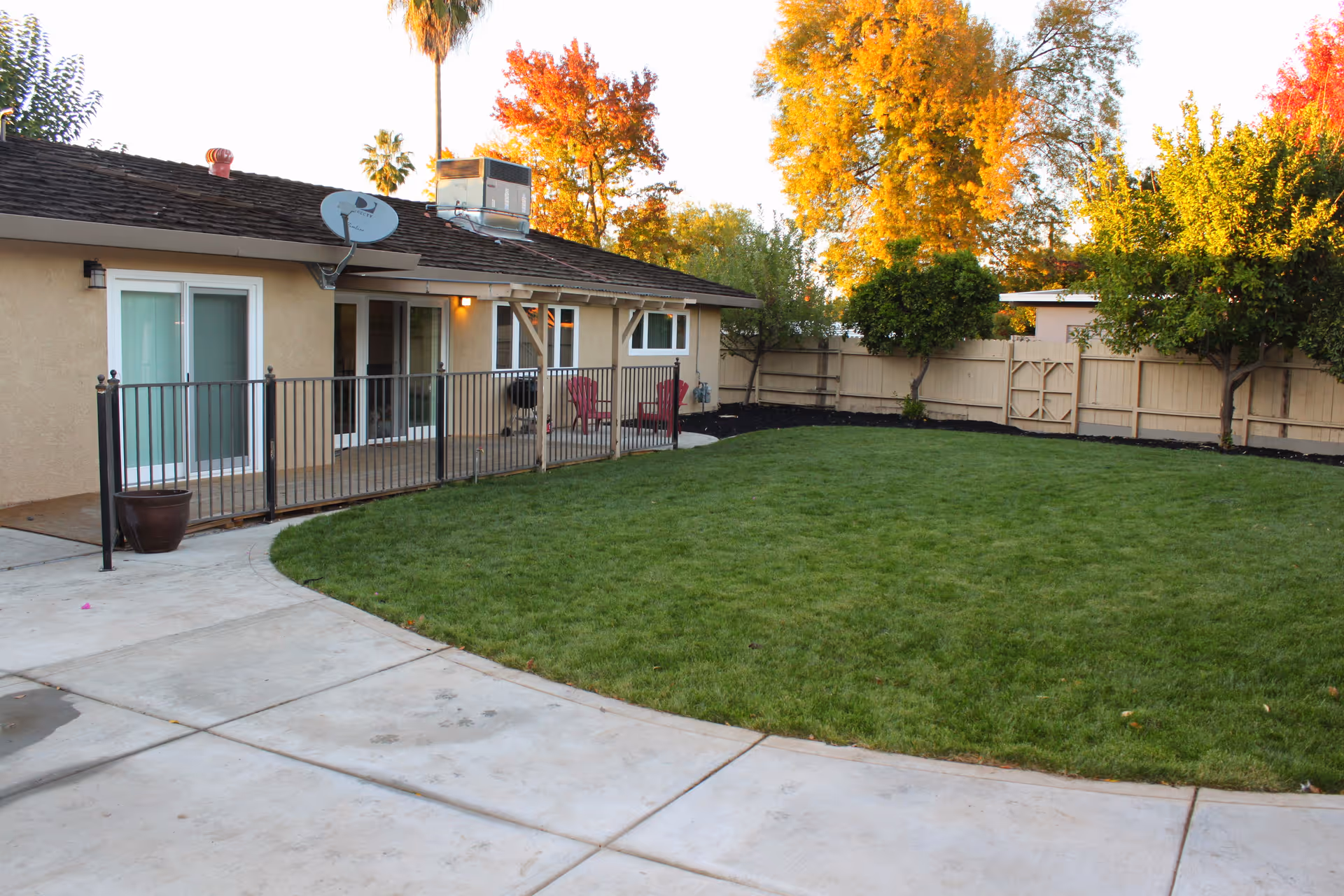 Backyard of a single-story building with a green lawn, concrete patio, fenced yard and sliding glass doors leading inside.