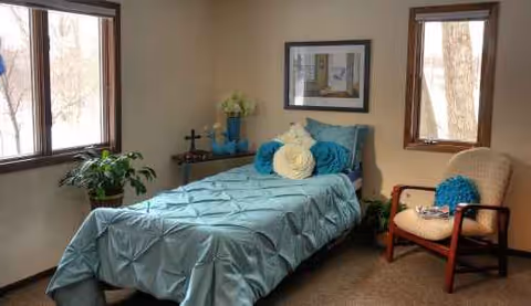 A small bedroom with teal bedspread, plants, and a cozy chair in a senior living facility.