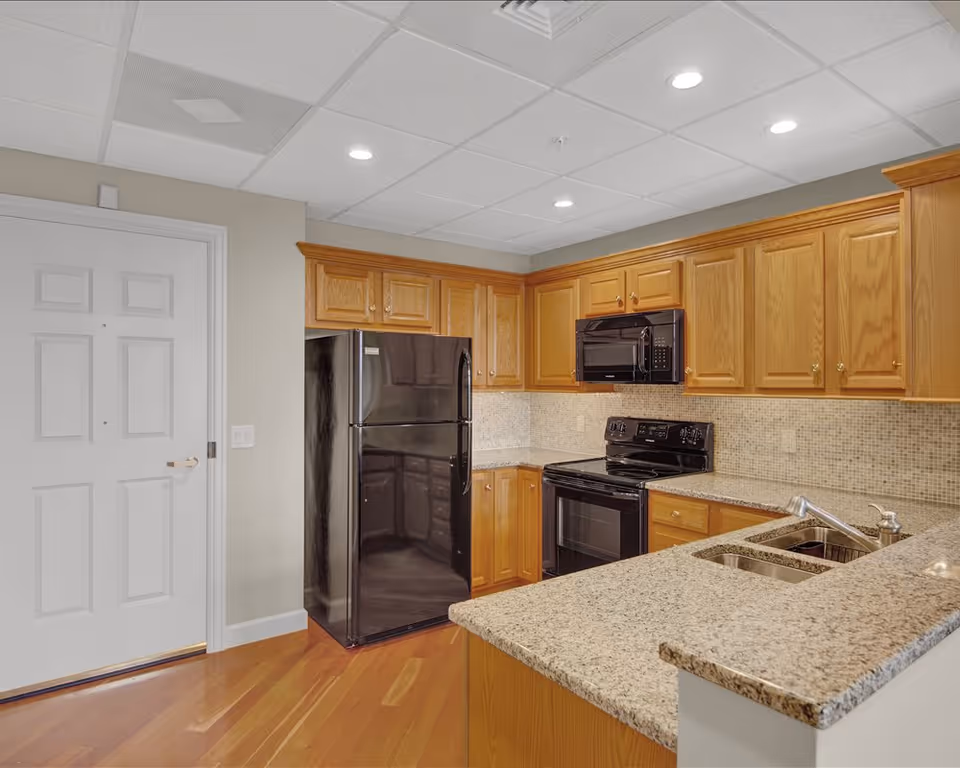 Interior view of a kitchen in a senior living community featuring wooden cabinets, a black refrigerator, black microwave, black stove, granite countertops, a double sink, and hardwood flooring. The ceiling has recessed lighting and a white door is visible on the left side.
