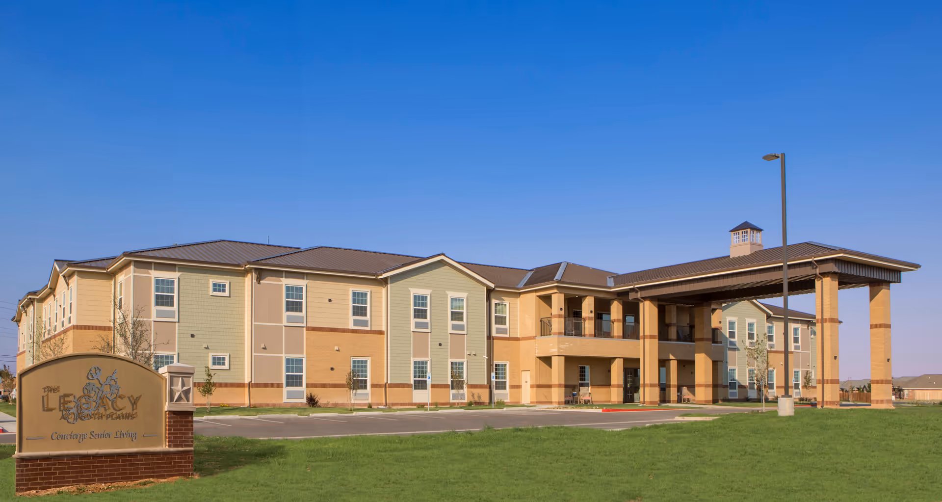 Exterior view of The Legacy at South Plains senior living facility showing a two-story building with a covered entrance, multiple windows, and a sign in front on a grassy lawn under a clear blue sky.