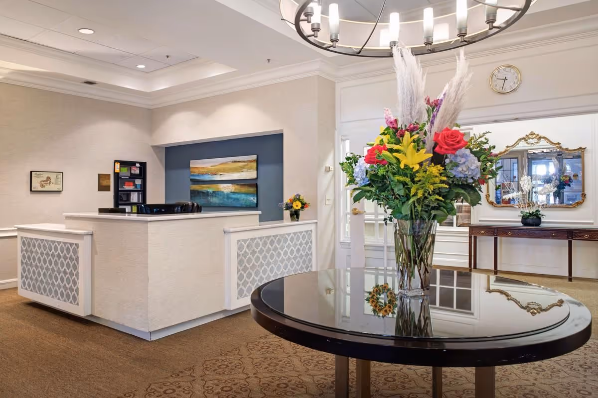 Reception area of Brighton Gardens of Friendship Heights featuring a white reception desk with patterned panels, a round table with a large colorful floral arrangement, a wall clock, a decorative mirror above a wooden console table, and a two-panel landscape painting on the wall behind the desk.