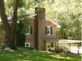 A brick residential building partially obscured by large trees with green leaves. The building has a prominent chimney, white-framed windows with black shutters, and a covered porch area with outdoor seating. The surrounding area is grassy and shaded by trees.
