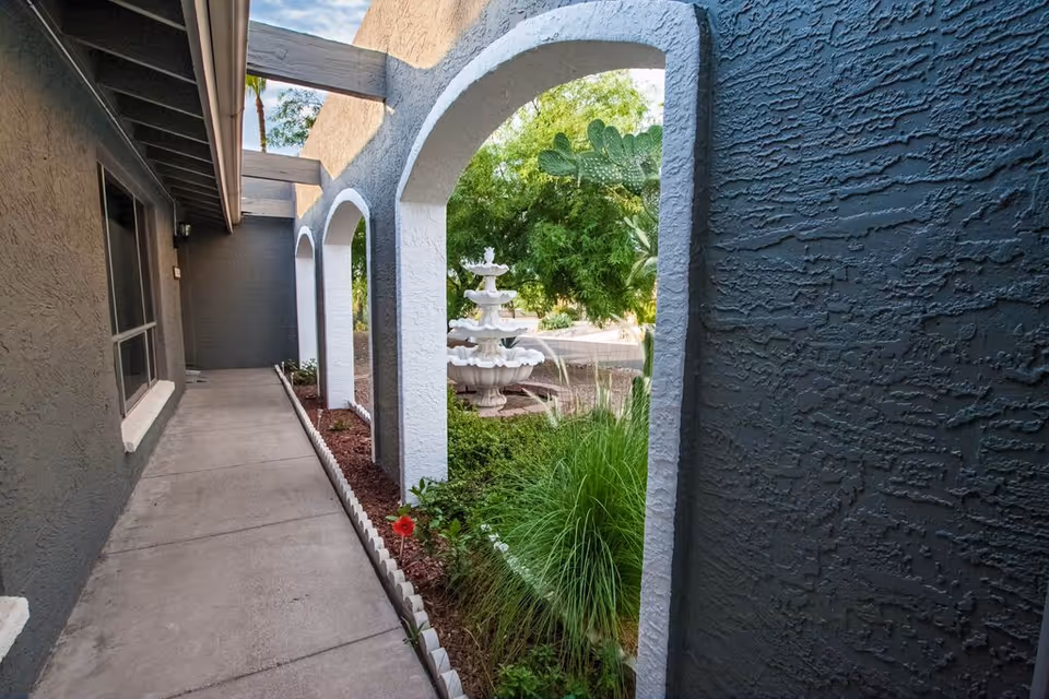 Outdoor walkway with a concrete path alongside a building with gray textured walls and white arched openings. Through the arches, a garden area with green plants and a white multi-tiered fountain is visible under a partly cloudy sky.